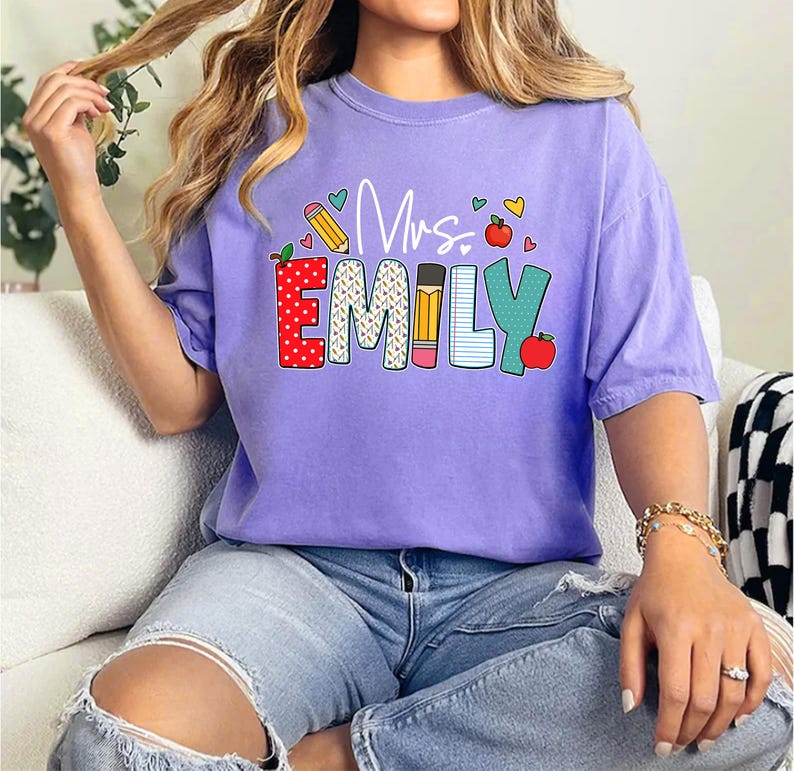 May include: Purple t-shirt with the text "Mrs. Emily" in colorful letters with a pencil, apple, and hearts design.