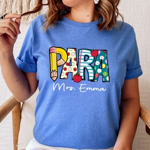 以下が含まれることがあります： 鉛筆、リンゴ、ストライプでカラフルな文字で「PARA」と綴られた青いTシャツ。 「Mrs. Emma」という文字が「PARA」の下に印刷されています。