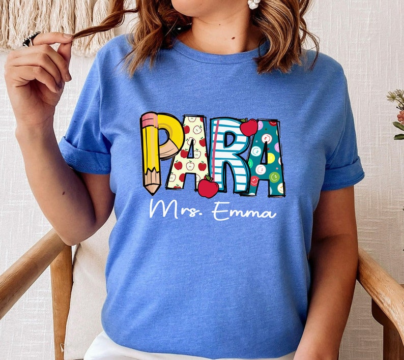 以下が含まれることがあります： 鉛筆、リンゴ、ストライプでカラフルな文字で「PARA」と綴られた青いTシャツ。 「Mrs. Emma」という文字が「PARA」の下に印刷されています。