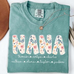 May include: A seafoam green Comfort Colors t-shirt with the word "NANA" in large, floral-filled letters. Below the word are names, each with a heart symbol. A silver necklace with a pendant rests on the shirt.