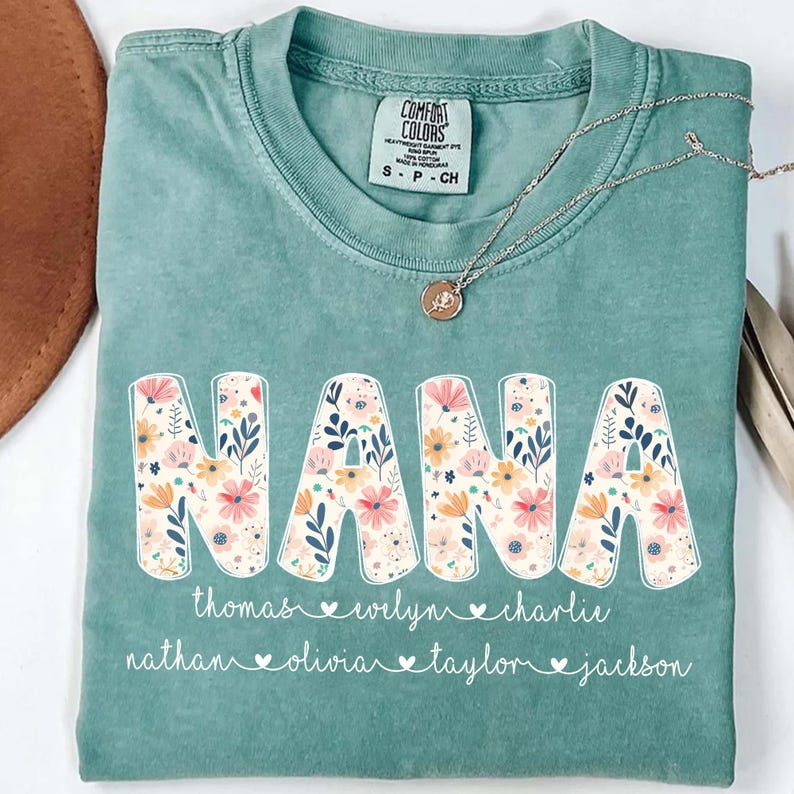 May include: A seafoam green Comfort Colors t-shirt with the word "NANA" in large, floral-filled letters. Below the word are names, each with a heart symbol. A silver necklace with a pendant rests on the shirt.