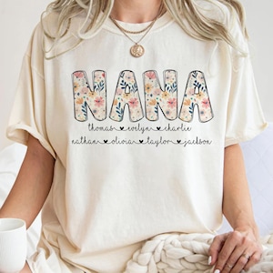 May include: Cream-colored t-shirt with the word "NANA" in large, floral-patterned letters. Below the word are names: Thomas, Evelyn, Charlie, Nathan, Olivia, Taylor, and Jackson. The shirt has a relaxed fit.