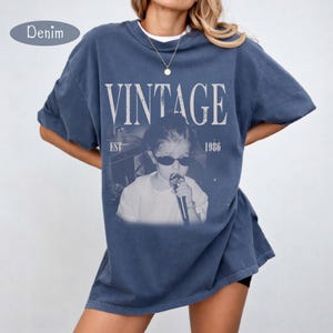 May include: A blue denim-colored t-shirt featuring a graphic print of a person wearing sunglasses and holding a microphone. The text VINTAGE is printed in large white letters above the image, with EST 1986 printed on the sides of the graphic.