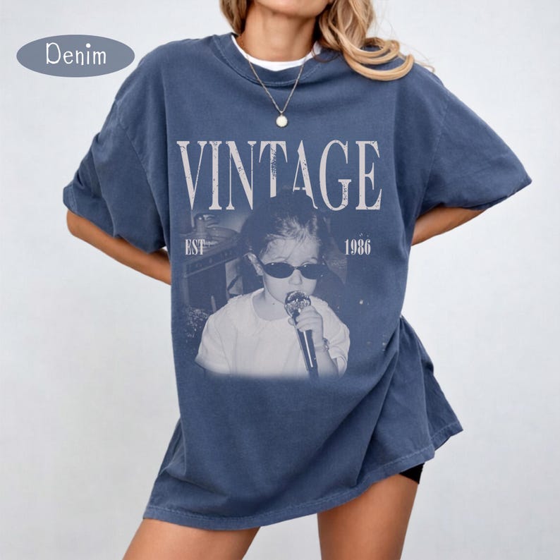 May include: A blue denim-colored t-shirt featuring a graphic print of a person wearing sunglasses and holding a microphone. The text VINTAGE is printed in large white letters above the image, with EST 1986 printed on the sides of the graphic.