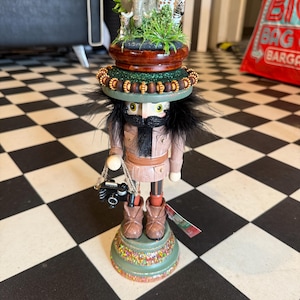 May include: A nutcracker figure dressed in a tan safari outfit with a black beard and hair. The hat is topped with miniature plastic safari animals including an elephant, giraffe, and zebra. The figure holds a small black camera on a rope.