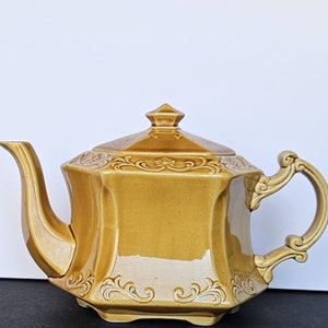 Peut inclure: Une théière en céramique brune avec un design orné et un corps carré. La théière a un couvercle avec un bouton sur le dessus et une poignée sur le côté.