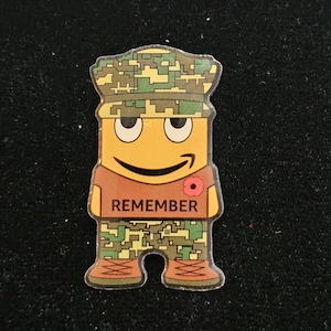 Peut inclure: Un autocollant plat découpé représentant un personnage de dessin animé portant une casquette et un pantalon à motif camouflage numérique, un t-shirt marron avec le mot REMEMBER imprimé en noir et une petite fleur de coquelicot rouge.