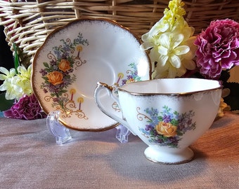 May include: A white teacup and saucer set with gold trim and floral designs. The cup and saucer feature a bouquet of orange roses, purple flowers, and green leaves. The set is displayed on a clear stand.