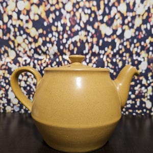 Könnte beinhalten: Eine keramische Teekanne in einem warmen Senfgelbton. Die Teekanne hat einen abgerundeten Körper, einen gebogenen Griff, einen Ausguss und einen Deckel mit einem kleinen Knopf. Der Hintergrund ist eine dunkle Oberfläche mit Bokeh-Effekt.