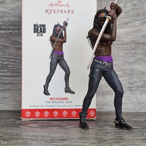 Puede incluir: Un adorno para árbol de Navidad Hallmark Keepsake de Michonne de The Walking Dead. La figura muestra un personaje con cabello largo oscuro, chaleco marrón, top morado y pantalones grises, sosteniendo una espada katana sobre la cabeza. Caja incluida.