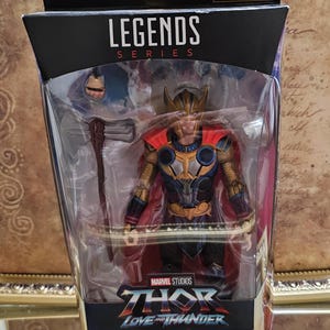 May include: A Marvel Legends Series action figure of Thor from Thor: Love and Thunder, packaged in a window box. The figure wears a gold, blue, and red suit with a red cape and includes a Stormbreaker axe accessory. The box features the movie logo.