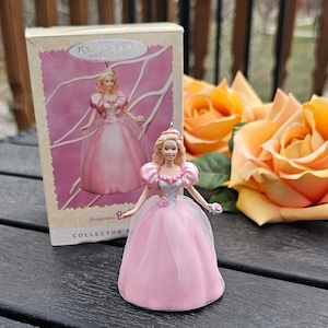 Peut inclure: Ornement de poupée Barbie rose avec une jupe en tulle et un diadème, à côté de sa boîte. La boîte présente une image correspondante et les mots "Keepsake Ornament" et "Springtime Princess Collector's Edition."