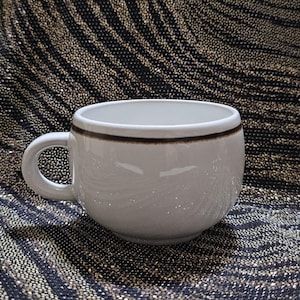 May include: A white ceramic coffee cup with a rounded shape and a handle. The cup has a brown band around the rim. The cup is sitting on a black and gold patterned surface.