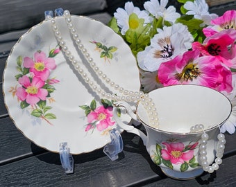 Puede incluir: Un juego de taza y platillo blancos con un diseño floral con flores rosas y hojas verdes. Un collar de perlas cuelga sobre el juego. La taza y el platillo están sobre una superficie oscura, con un ramo de flores rosas y blancas en el fondo.