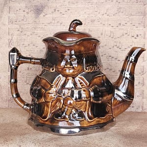 May include: A brown ceramic teapot with a lid, handle, and spout. The teapot is shaped like a man in a suit with silver accents. The teapot has a glossy finish and is set against a background of sheet music.