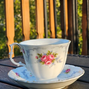 Puede incluir: Un juego de taza y platillo blancos con un diseño floral con rosas rosas y detalles azules. La taza tiene un asa curvada y ribete dorado. El platillo refleja el diseño de la taza. El juego está sobre una superficie de madera oscura.