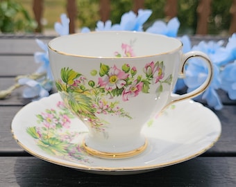 Puede incluir: Una taza de té y un platillo blancos con un patrón floral con flores rosas y verdes. La taza y el platillo tienen un borde dorado.