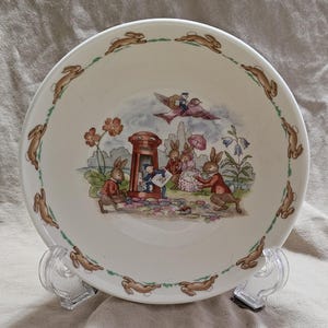 May include: A white ceramic bowl with a charming illustration of rabbits, a postbox, and a bird carrying a parcel. The rim is adorned with running rabbit figures. The bowl is supported by a clear plastic stand.