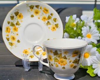 May include: White teacup and saucer set with a floral pattern in yellow and green. The teacup has a rounded base and a curved handle. The saucer is round with a matching floral design. Both pieces have gold trim.