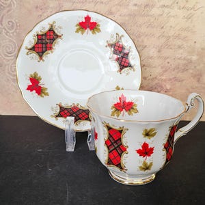Peut inclure: Une tasse à thé et une soucoupe blanches avec un motif de tartan rouge et noir et des accents de feuilles d'érable. L'ensemble est orné d'une bordure dorée.