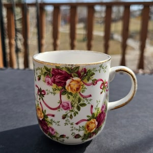 Può includere: Tazza in ceramica bianca con un motivo floreale di rose gialle, rosa e bordeaux, accentuato da foglie verdi e nastri rosa. Il bordo e il manico sono rifiniti in oro. Una tazza decorativa per bevande.