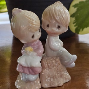 Könnte beinhalten: Porzellanfiguren-Set mit einem Jungen und einem Mädchen. Das Mädchen trägt ein rosa Kleid und hält eine gelbe Blume. Der Junge sitzt auf einem Baumstumpf. Beide Figuren haben einen ruhigen Ausdruck und sind hell gehalten.