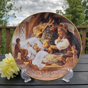 Puede incluir: Plato decorativo con una ilustración detallada de una familia de osos en un dormitorio. El plato tiene un borde dorado y se exhibe en un soporte de plástico transparente. La escena incluye un niño en la cama, un oso de peluche y otras figuras.