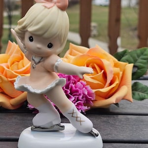 May include: A porcelain figurine of a skater with blonde hair and a peach bow, wearing a peach dress with white trim, white socks, and white skates with gold cross-lacing details. The figure stands on a round white base with a silver-toned necklace.