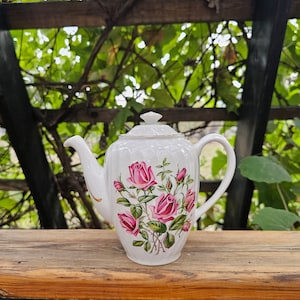 May include: A white porcelain teapot featuring a pink rose floral pattern. The teapot has a curved spout, a handle, and a lid with a decorative finial. Gold trim accents the edges. The teapot is displayed on a wooden surface with a green leafy background.