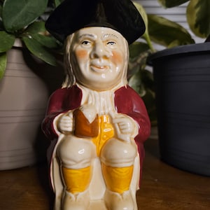 Może przedstawiać: Ceramiczna figurka dzbanka Toby przedstawiająca siedzącego mężczyznę w trójgraniastym kapeluszu i czerwonym płaszczu, trzymającego kufel i butelkę. Postać ma uśmiechniętą twarz z różowymi policzkami i nosi żółte bryczesy i białe buty. Dzbanek to przedmiot dekoracyjny.