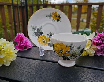 May include: A white teacup and saucer set with a floral design featuring yellow and gray flowers. The cup has a footed base and a gold rim. The saucer also has a gold rim. The set is displayed on a black surface with colorful flowers in the background.