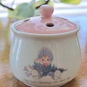 May include: A white ceramic jar with a pink lid and a small illustration of a child holding a Christmas tree. The jar has the text "May God's Blessings Fall Upon You" on the bottom.