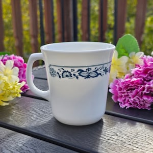 Puede incluir: Taza de cerámica blanca con un diseño floral azul alrededor del borde. La taza tiene un asa curvada y está colocada sobre una superficie gris oscuro, con flores coloridas en el fondo. La taza está vacía.