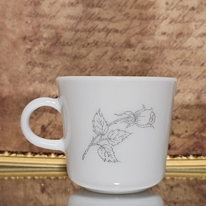 Könnte beinhalten: Weiße Keramik-Tasse mit gebogenem Henkel. Eine graue Punktzeichnung einer Rose mit Blättern verziert die Vorderseite. Die Tasse steht auf einer reflektierenden Oberfläche.