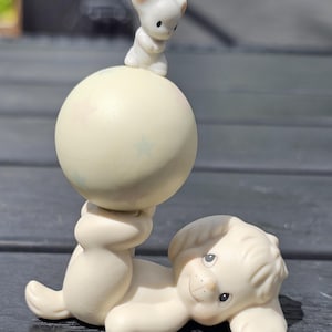 Könnte beinhalten: Eine matte, cremefarbene Porzellanfigur eines Welpen, der auf der Seite liegt und eine große, blassgelbe Kugel auf seinen Hinterpfoten balanciert. Eine kleine weiße Maus sitzt auf der Kugel, die mit blassen, hellblauen Sternenmustern verziert ist.