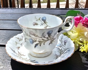 Può includere: Una tazza da tè e un piattino bianchi con un motivo floreale che presenta cardi blu e foglie verdi. La tazza e il piattino hanno un bordo dorato.