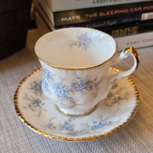 Puede incluir: Un juego de taza y platillo blancos con un estampado floral azul y ribete dorado. La taza tiene un asa curvada y se apoya en un platillo a juego. El juego está colocado sobre una superficie clara.