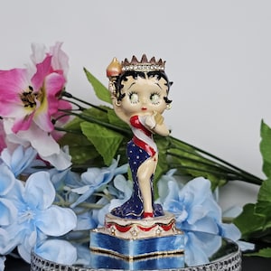 May include: A ceramic figurine of Betty Boop dressed as the Statue of Liberty. She is wearing a blue and white dress with red stripes and a crown. She is holding a torch in her right hand and a book in her left hand.