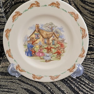 May include: A cream-colored ceramic plate featuring a charming illustration of rabbits dancing around a house. The plate's border is adorned with running rabbit figures and green grass accents, creating a whimsical, storybook aesthetic.