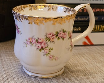May include: A white porcelain teacup with a gold rim and handle, decorated with pink rose floral patterns. The cup features a wide base and a curved handle. The rim and handle are trimmed with gold filigree.