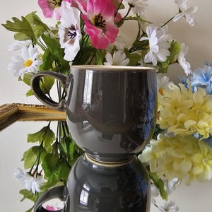 Op de afbeelding: Een donkergrijze keramische mok met een brede rand en een handvat. De mok wordt weerspiegeld in een spiegel met een gouden lijst. De mok is omringd door een boeket roze, witte en gele bloemen.