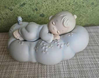 Może przedstawiać: Porcelanowa figurka śpiącego dziecka na chmurze z małym ptakiem siedzącym na krawędzi. Dziecko ma na sobie biały śpioszek i ma spokojny wyraz twarzy. Chmura jest ozdobiona małymi różowymi kwiatami.