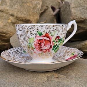 May include: A vintage teacup and saucer set. The white porcelain cup and saucer are decorated with a red rose and gold floral pattern. The teacup has a curved handle. The set is trimmed with gold detailing, adding to its classic appeal.