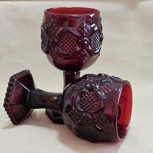 Puede incluir: Dos copas de color rojo rubí con intrincados patrones florales y geométricos en relieve. Una copa está en posición vertical, la otra está de lado. Las copas tienen un tallo texturizado y una base festoneada. El cristal tiene un rico color rojo oscuro.