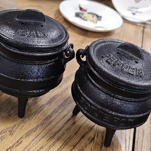 Peut inclure: Deux salières et poivrières en fonte noire, chacune avec un couvercle et trois pieds. La salière porte l'inscription "SALT" sur le couvercle, et la poivrière porte l'inscription "PEPPER". Les deux ont de petites poignées.