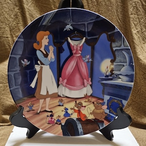 Puede incluir: Plato decorativo con una escena de Cenicienta. El plato representa a Cenicienta con un vestido azul y blanco, un vestido rosa y varios ratones. El fondo incluye un interior de castillo con chimenea y luna llena.