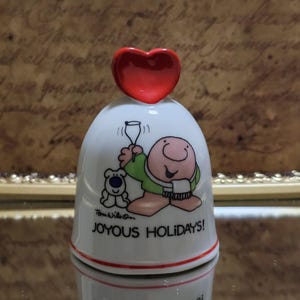 May include: A white ceramic bell with a red heart-shaped top and a cartoon illustration. The bell features a character holding a glass and a bear, with the text "JOYOUS HOLIDAYS!"
