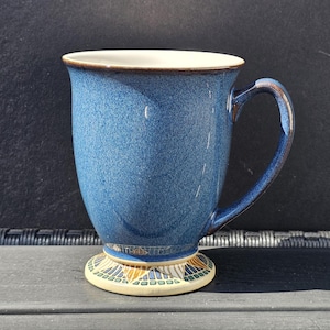 Op de afbeelding: Een keramische mok met een gespikkeld blauw glazuur en een gladde, crèmekleurige binnenkant. De voet is voorzien van een decoratief patroon van blauwe, groene en beige geometrische vormen. De mok heeft een gebogen handvat en een uitlopende rand.