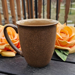 Op de afbeelding: Een bruine keramische koffiemok met een gespikkelde afwerking en een afgeronde handgreep. De mok staat op een donker houten oppervlak met oranje bloemen op de achtergrond.