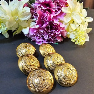 May include: Six gold-coloured metal buttons with a decorative design. Each button features a wreath with a crown and a fleur-de-lis in the centre.